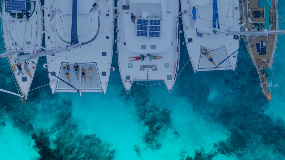 Catamarans anchored together over clear shallow water, people resting on the trampolines